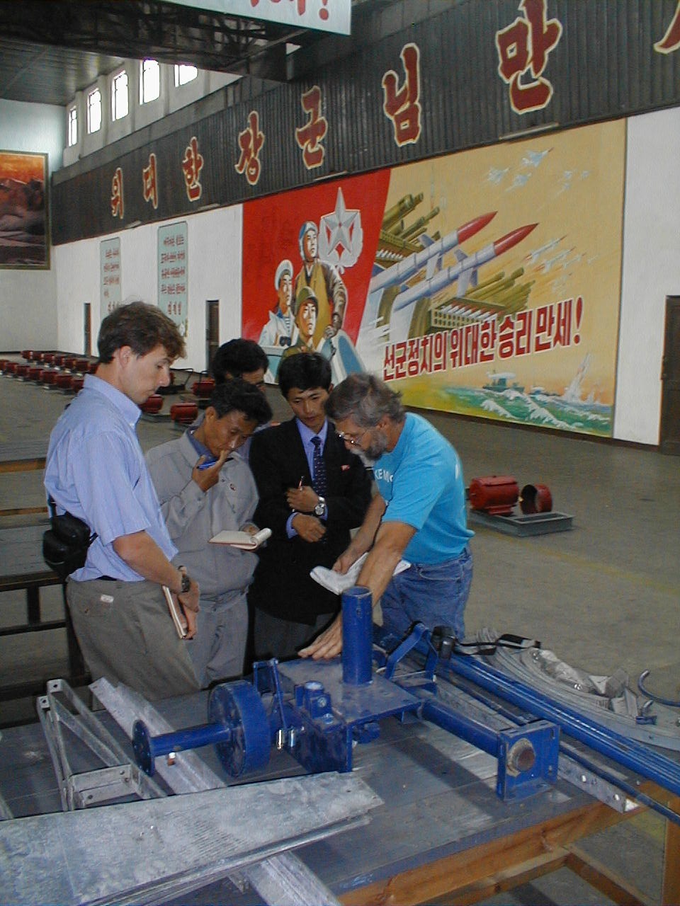 Nautilus team in Electric Appliance Factory #1 in Pyongyang inspecting wind turbines in 1998. On the wall behind, the big horizontal banner says, “Long Live the great general” (that is, Kim Jong-il).    The poster with missiles below that banner says, “Long Live the Great Victory of Seongun Jeongchi” (that is, Military First Politics) 
